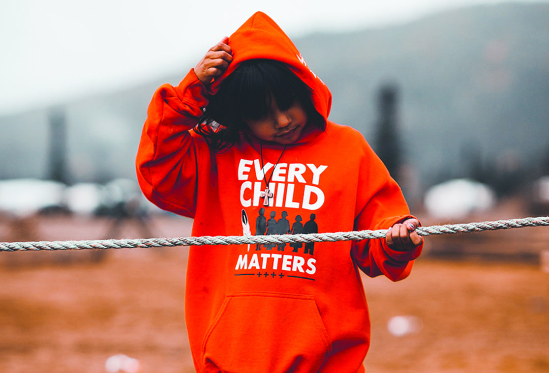 A young person wearing an orange hooded sweatshirt printed with text that reads Every Child Matters