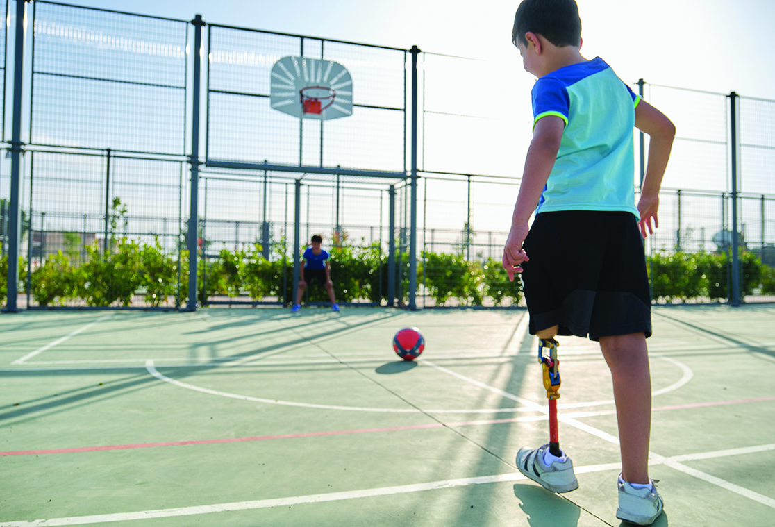 Two friends playing football, one of them has a leg prosthesis and is kicking a penalty. Kids playing sports together.