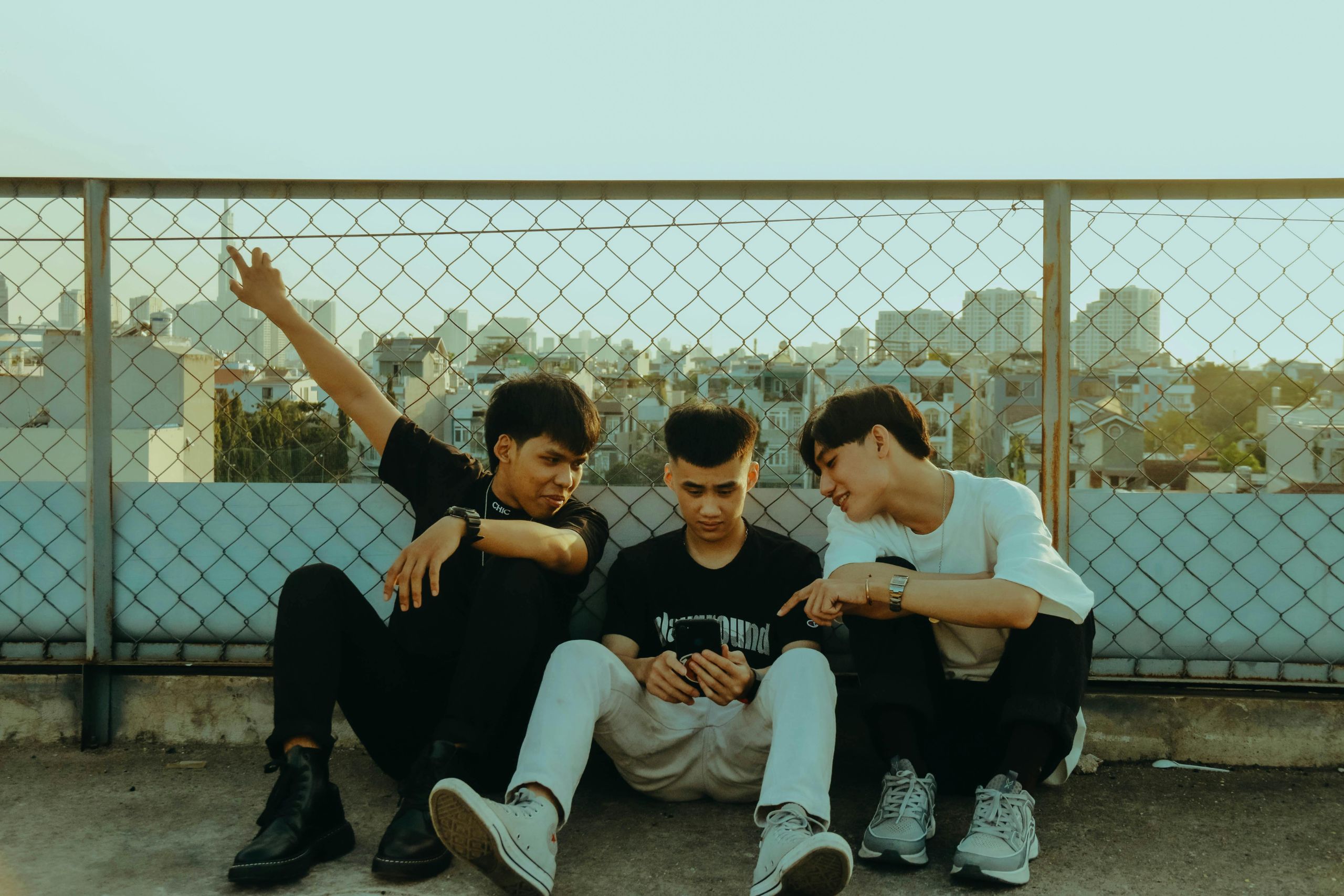 Three newcomer youth sitting together on a rooftop near a chain-link fence with a cityscape in the background
