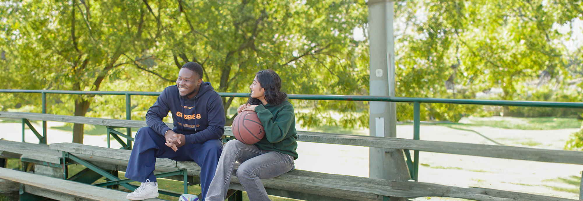 Two young people wearing KHP merch sitting on the bleachers with a basketball