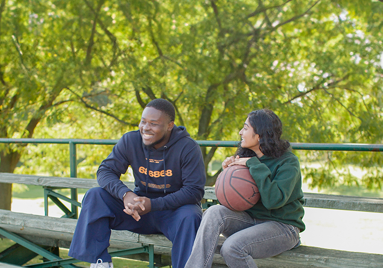 Two young people wearing KHP merch sitting on the bleachers with a basketball