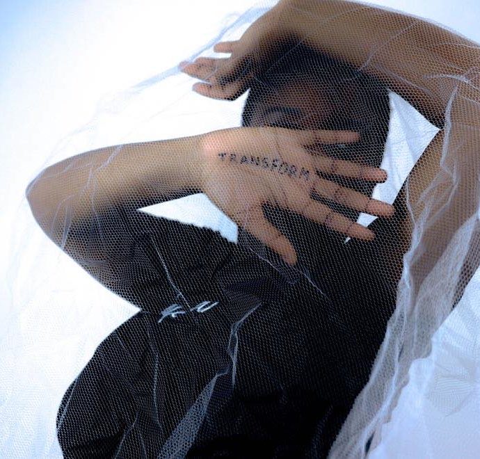 A self-portrait of someone with light netting over their body and “transform” written on their hand.